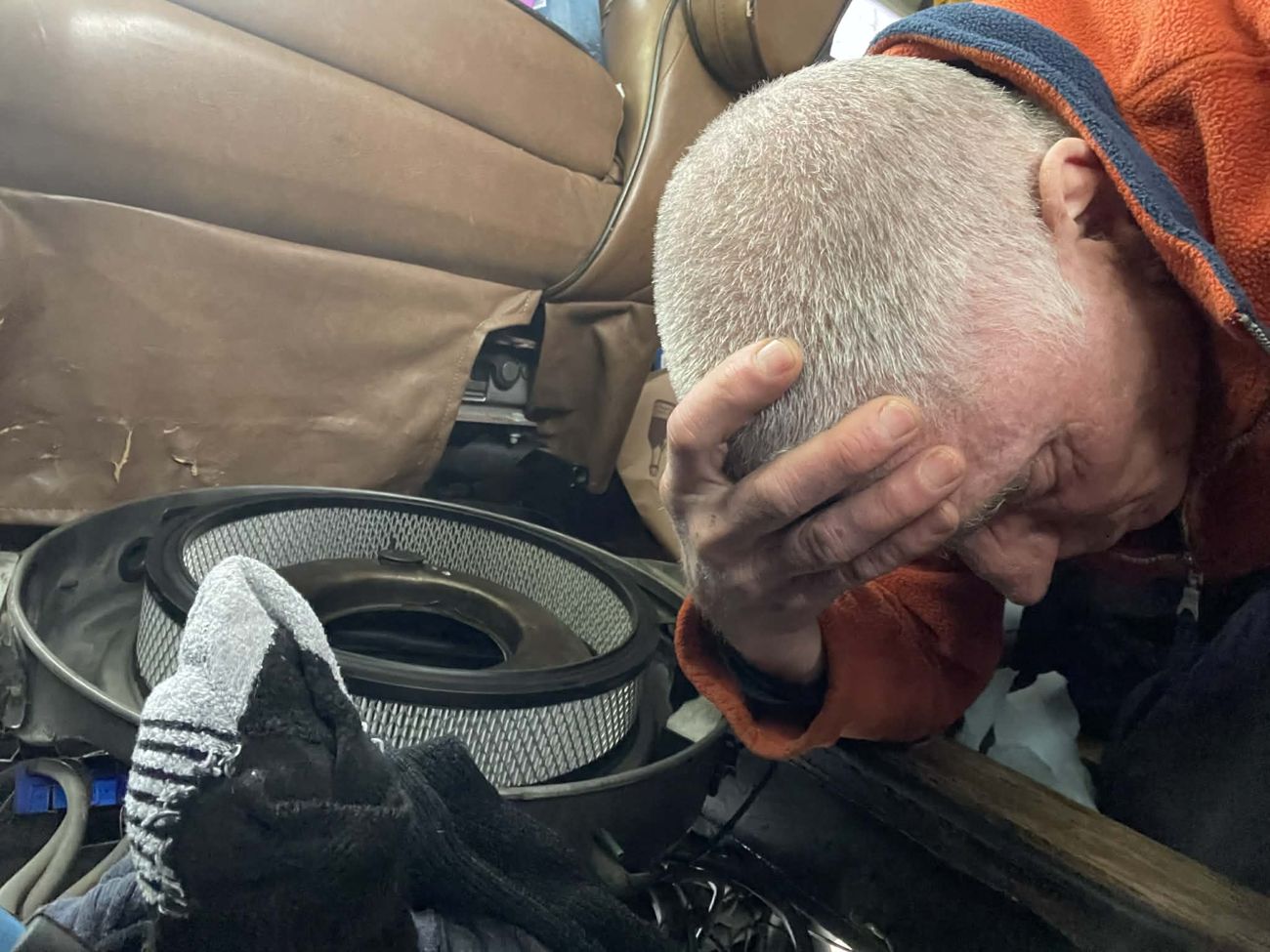 Close photograph of a man in an orange fleece with his hand pressed to his forehead, eyes shut, leaning over an air filter housing and a black glove resting in his lap inside a vehicle cab.