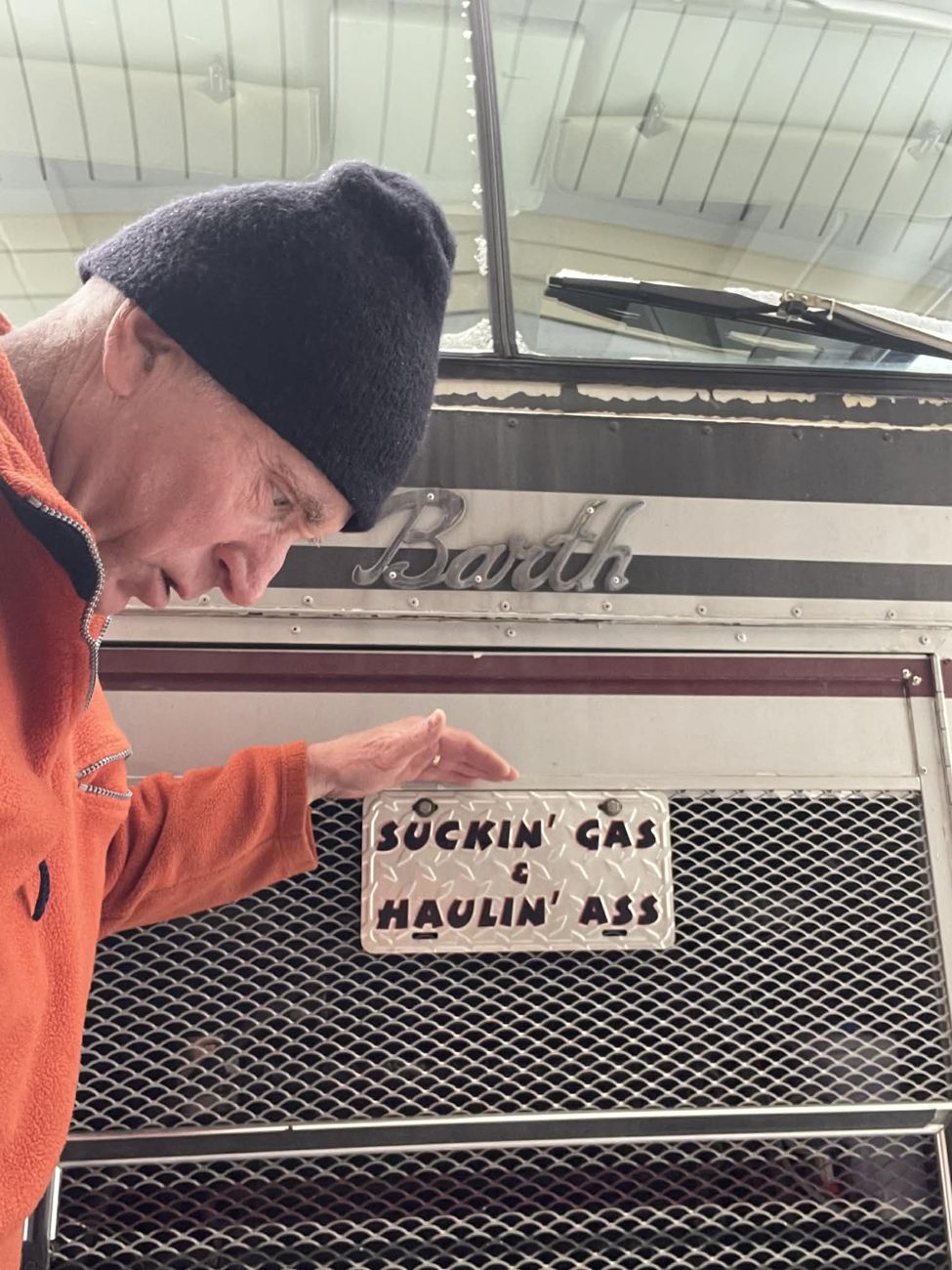 Photograph of a man in an orange fleece and dark beanie leaning in beside the front grille of the Barth, pointing at a white placard mounted on the mesh that reads SUCKIN' GAS AND HAULIN' ASS in hand-painted capitals. The chrome Barth script is visible above.
