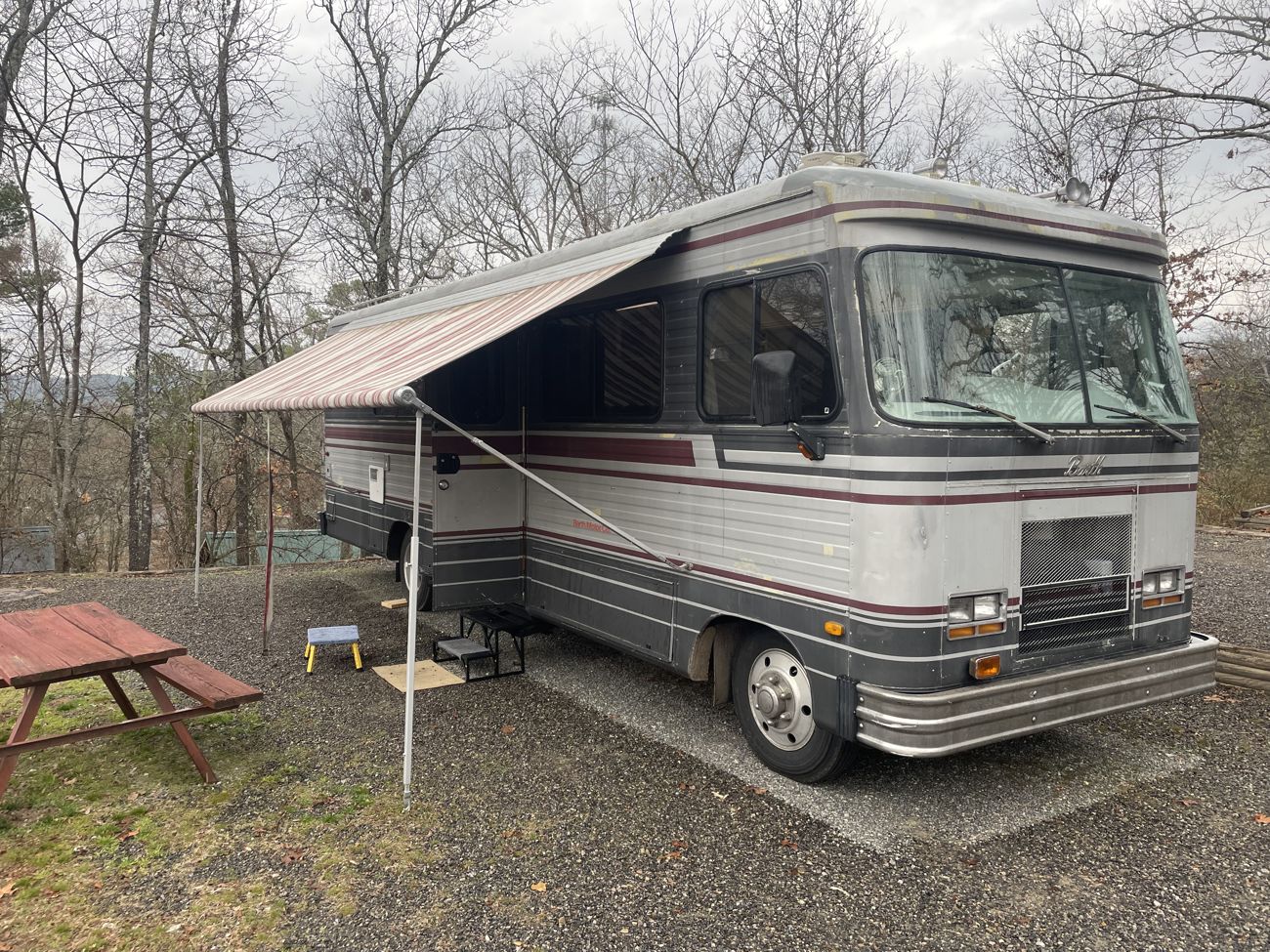 Photograph of the Barth motorhome parked on a gravel campsite pad among bare winter trees, its main awning extended over a small step stool and folding table, a wooden picnic table in the foreground. Overcast sky, dry leaves on the ground.