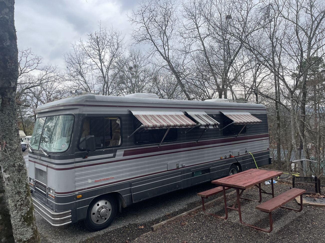 Photograph of the Barth motorhome at a wooded campsite with its main awning and all three window awnings deployed, picnic tables in the foreground, bare trees and a bright cloudy sky behind.