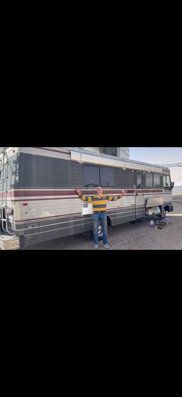 Photograph of a man in a yellow and blue striped rugby shirt standing with both arms spread wide in front of the long side of the Barth motorhome on open ground, engine hatch open behind him, flat scrubland and grey sky beyond.