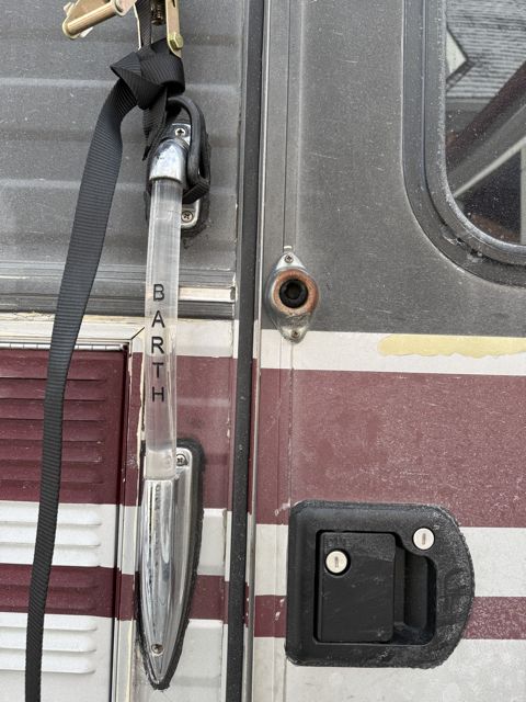 Close photograph of the side of the motorhome showing a black ratchet strap with a brass mechanism pulled tight over a chrome metal remnant, the vertical chrome BARTH lettering below it, a black door latch to the right and the maroon stripe crossing the frame.