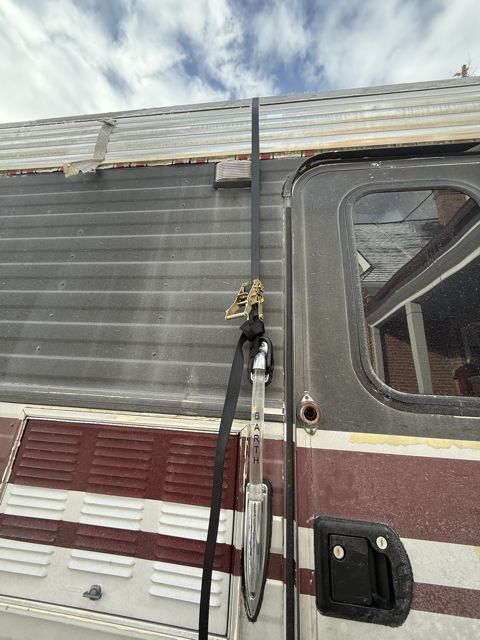 Photograph of the motorhome's upper side panel showing the empty awning track along the roofline where the awning was cut away, a torn edge above the window, and the ratchet strap running down the body.