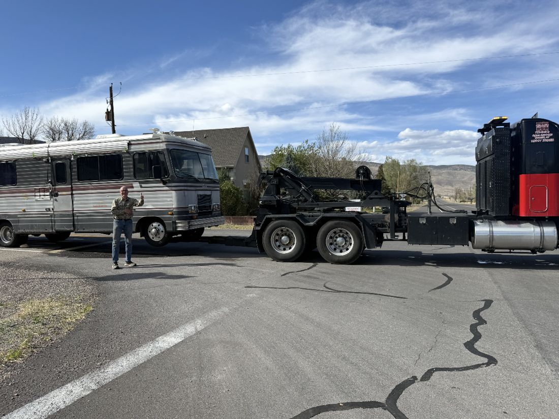 On a rural Utah roadside under a blue sky, the long grey Barth motorhome sits at the kerb with a man standing beside it, thumb raised, while a large red and black tow truck with its boom extended is positioned across the road ready to hook up. Houses and dry hills behind.